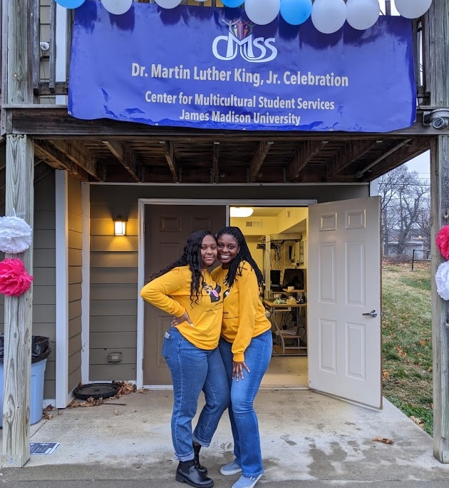 two student volunteers standing in front of the Reservoir St basement below a sign that reads JMU CMSS Martin Luther King Jr Celebration
