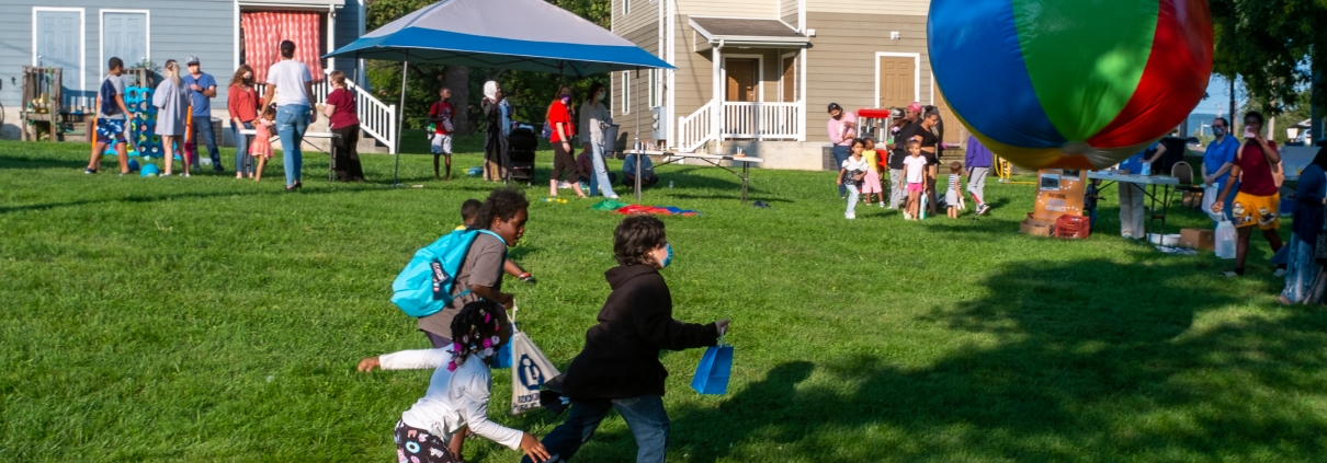 Children chase a large beach ball in the foreground as families mingle with community partners in the back.
