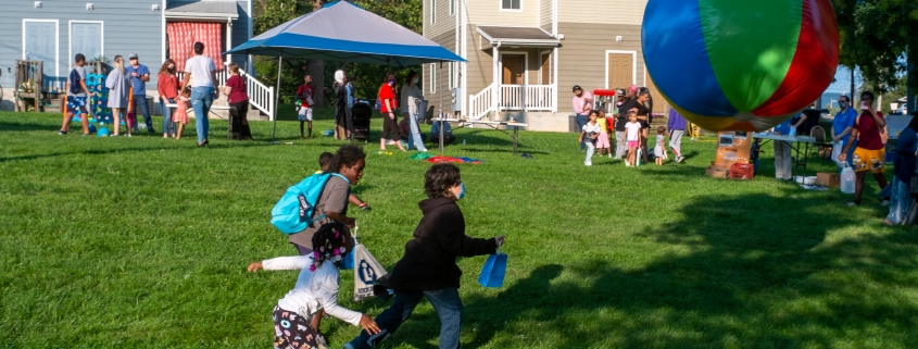 Children chase a large beach ball in the foreground as families mingle with community partners in the back.